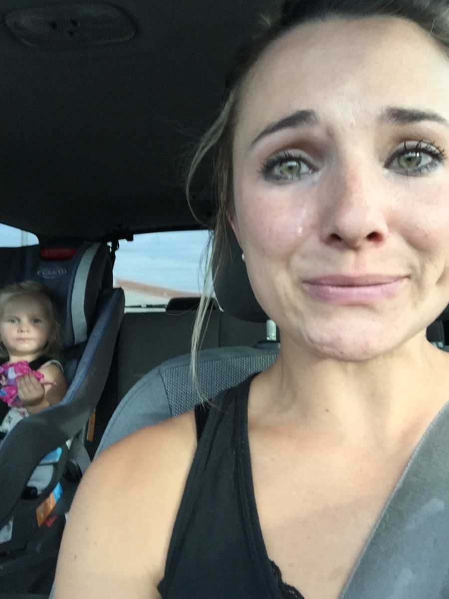 Mother cries in drivers seat of car while young daughter sits in backseat in carseat