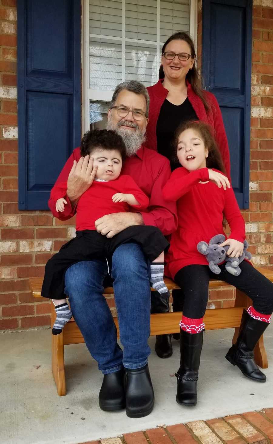 Man sits on bench outside of home with foster son and daughter while man's wife stands behind them