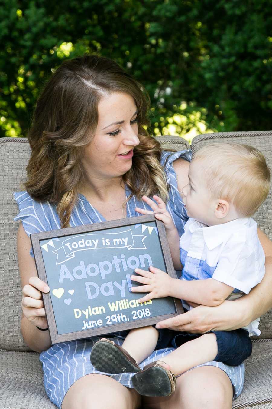 Mother sits outside on couch with foster son she is going to adopt on her lap along with sign announcing adoption