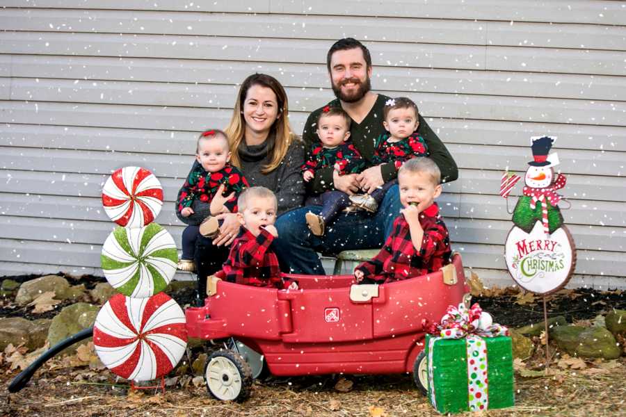 Husband and wife sit on bench holding triplets while their two adopted sons sit in red wagon beside Christmas decorations