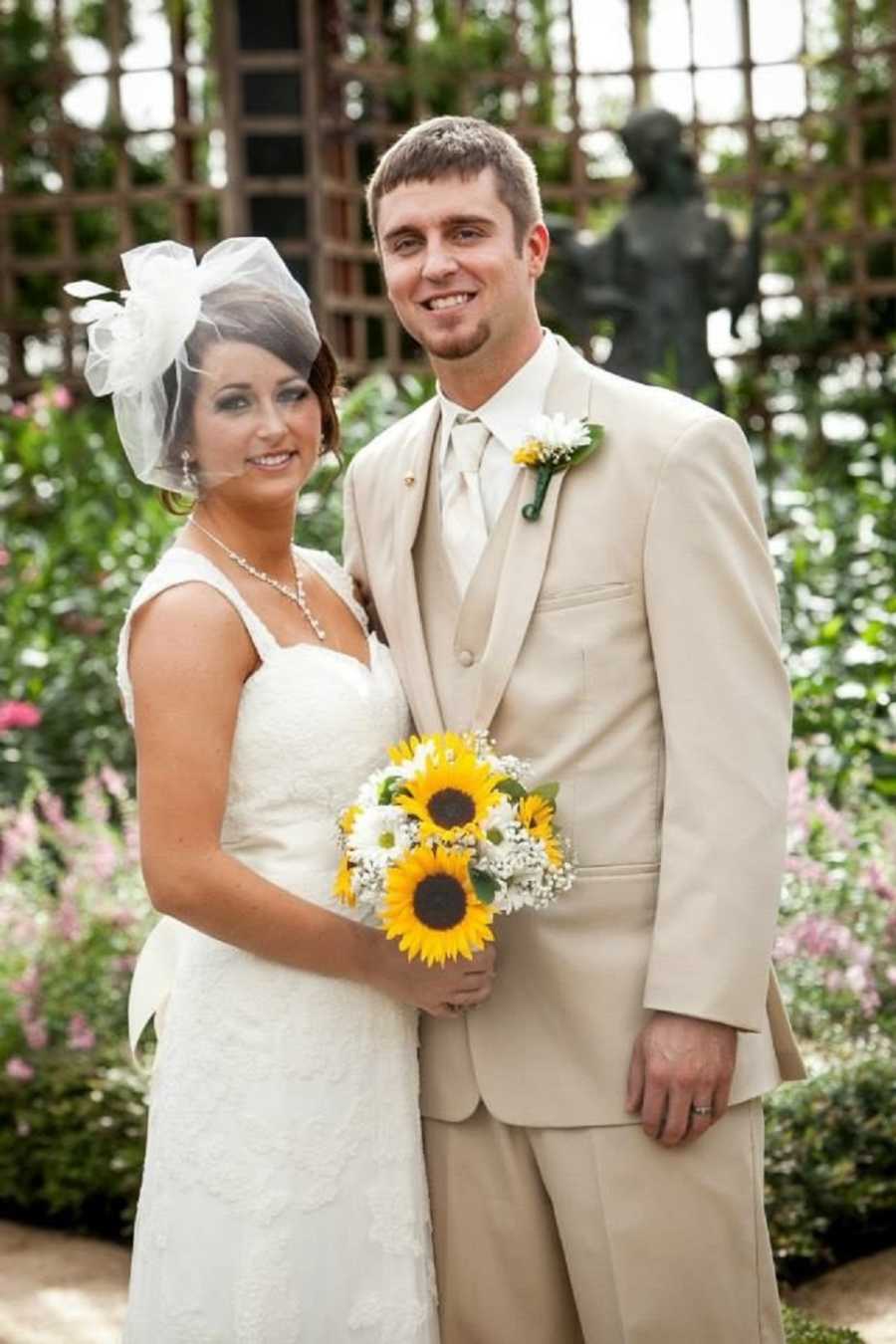 Bride stands holding bouquet of sunflowers outside beside groom