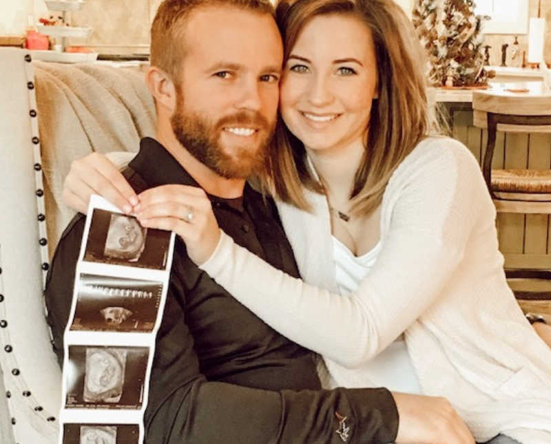 Wife sits on husbands lap in home holding ultrasound pictures