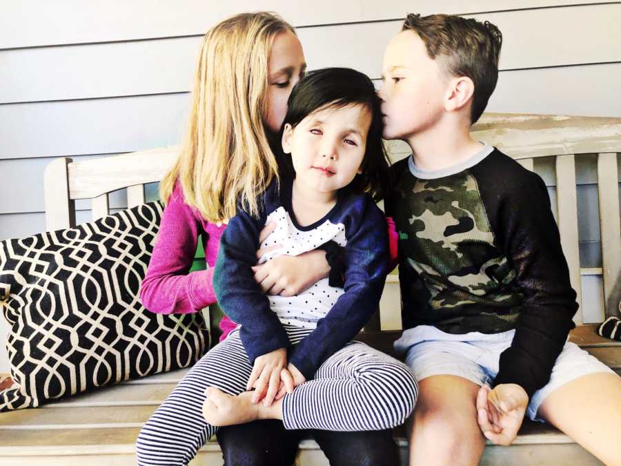 Adopted little girl sits on older sister's lap while she and brother kiss sister's head