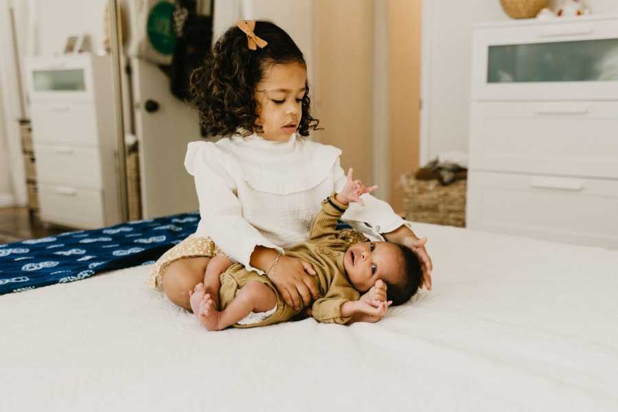 Little girl sits on bed with adopted baby sister laying beside her