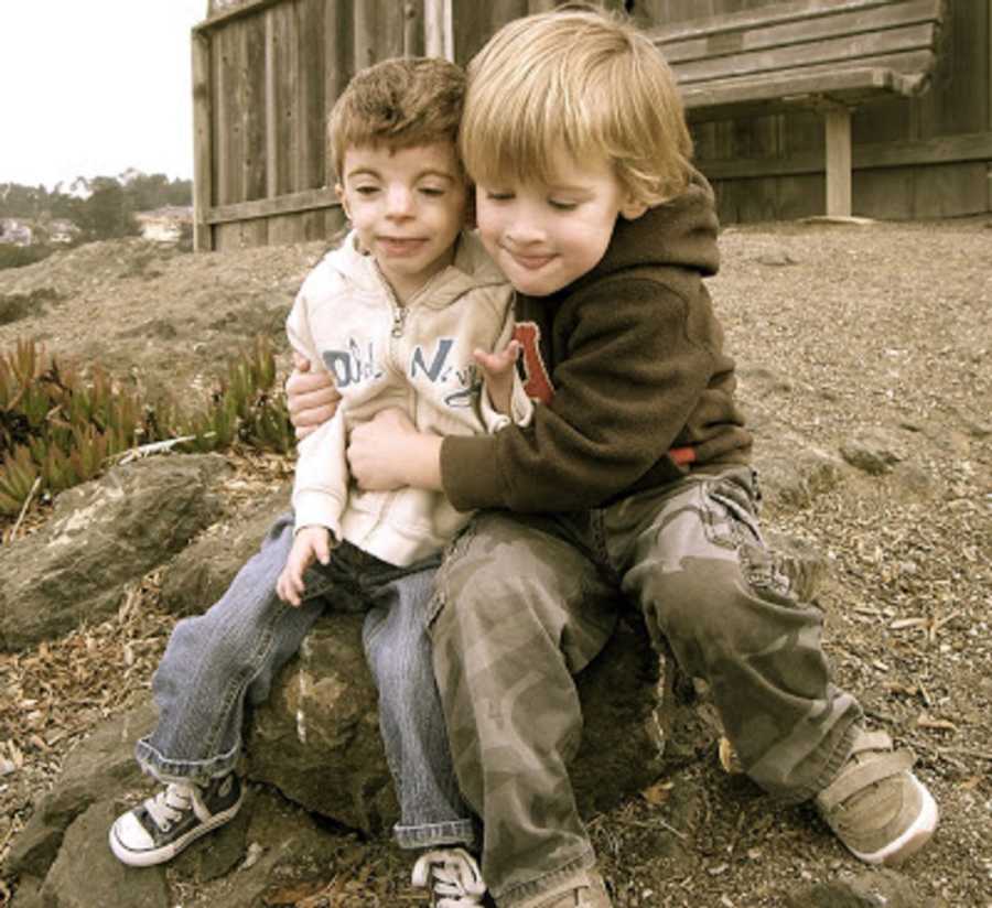 Young boy with Cornelia de Lange Syndrome sits on rock outside with brother who hugs him