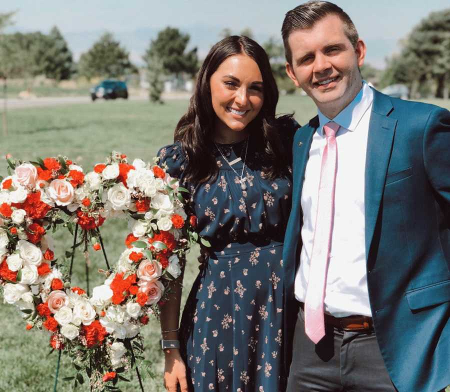 Husband and wife stand smiling outside beside bouquet of flowers shaped as heart