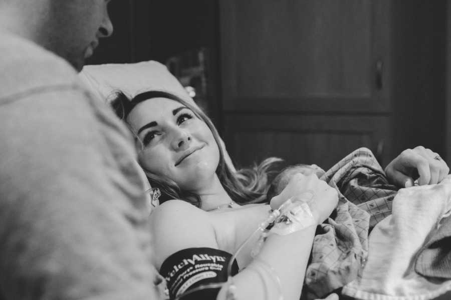 Woman lays in hospital bed with newborn on her chest as she smiles at her husband