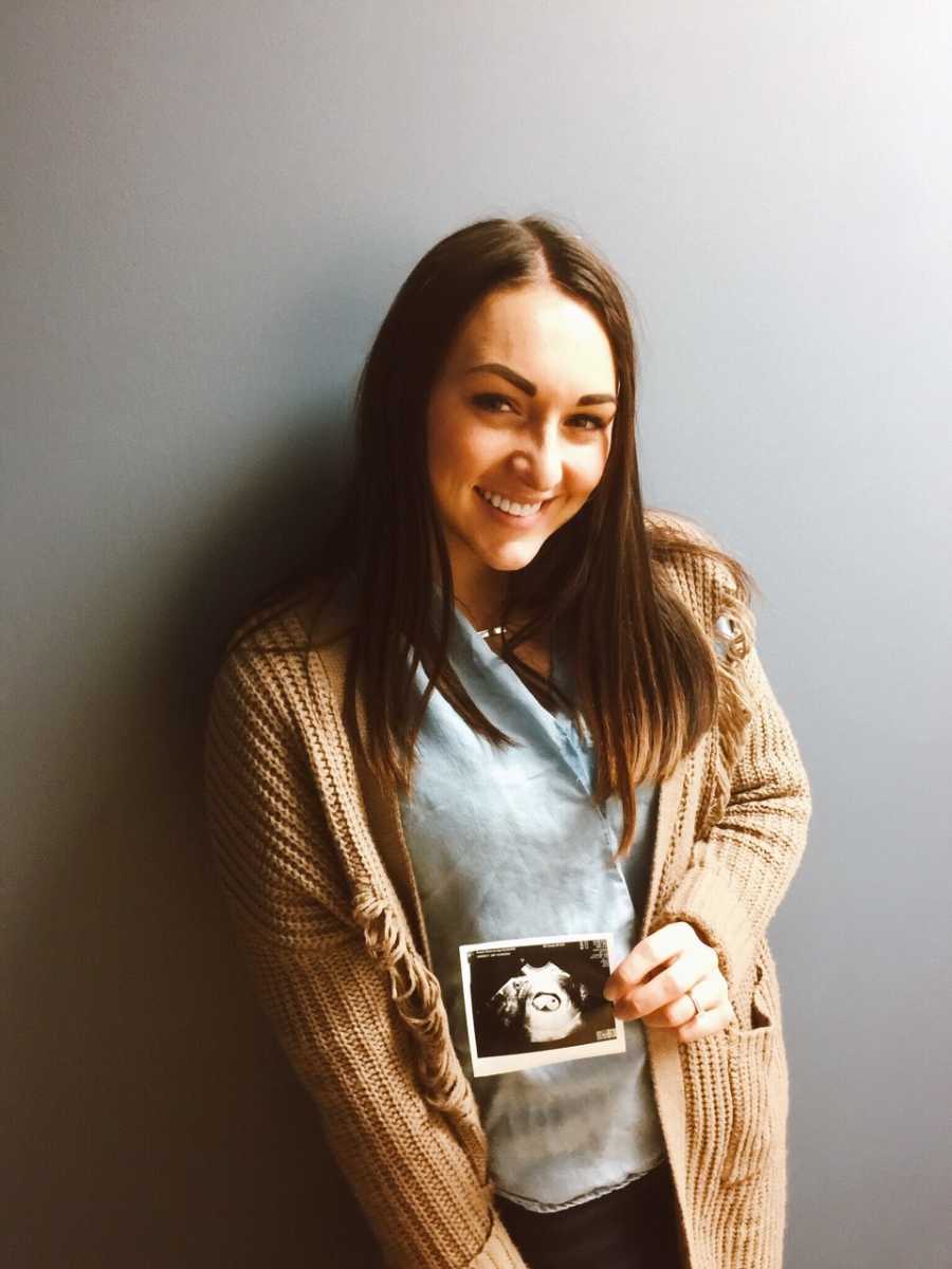 Woman stands smiling holding ultrasound picture in front of her stomach