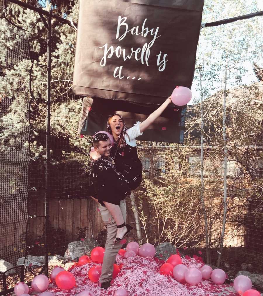 Husband stands outside holding wife at gender reveal with pink balloons and confetti at his feet