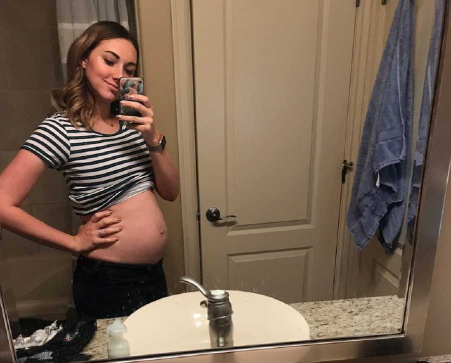Pregnant woman smiles in mirror selfie in her bathroom with shirt raised to show her stomach
