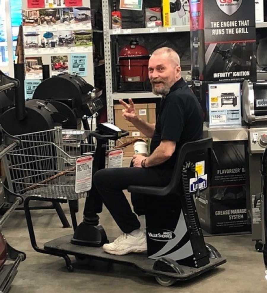 Man who has brain tumor sits in electric shopping cart holding up peace sign