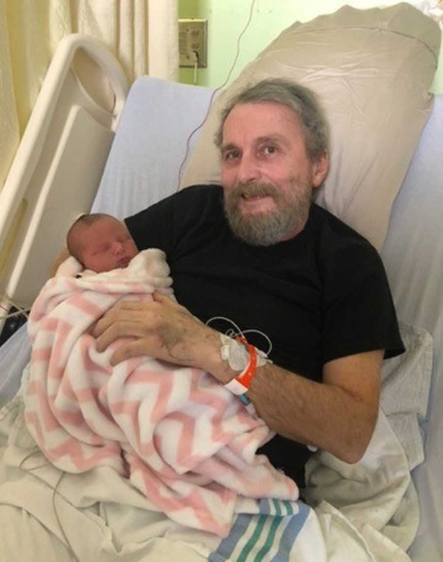 Man with brain tumor lays in hospital bed holding newborn granddaughter