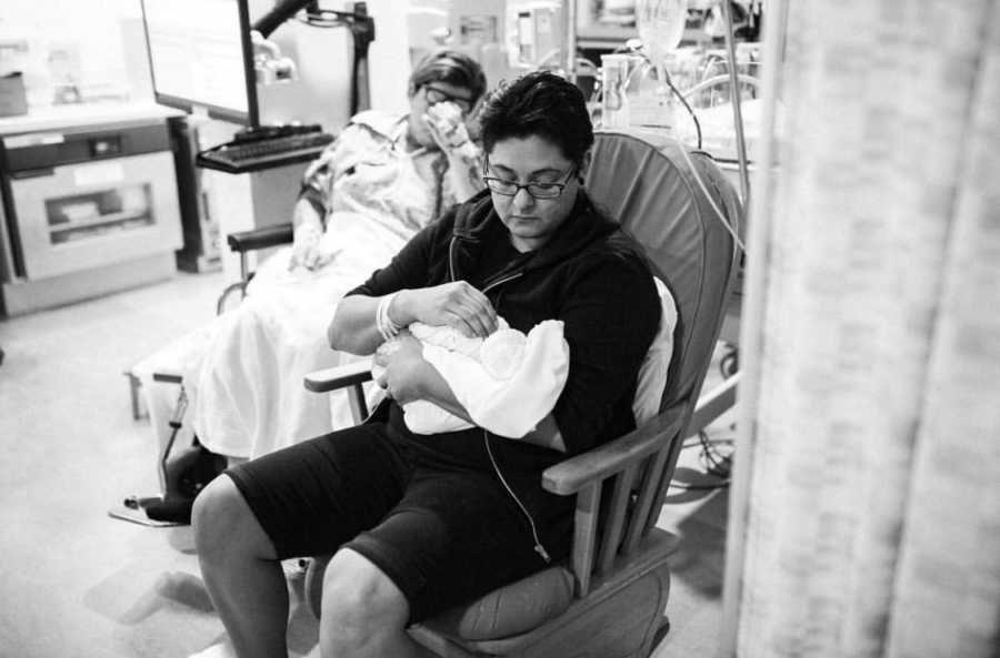 Mother sits in rocking chair in NICU holding newborn with wife in background crying