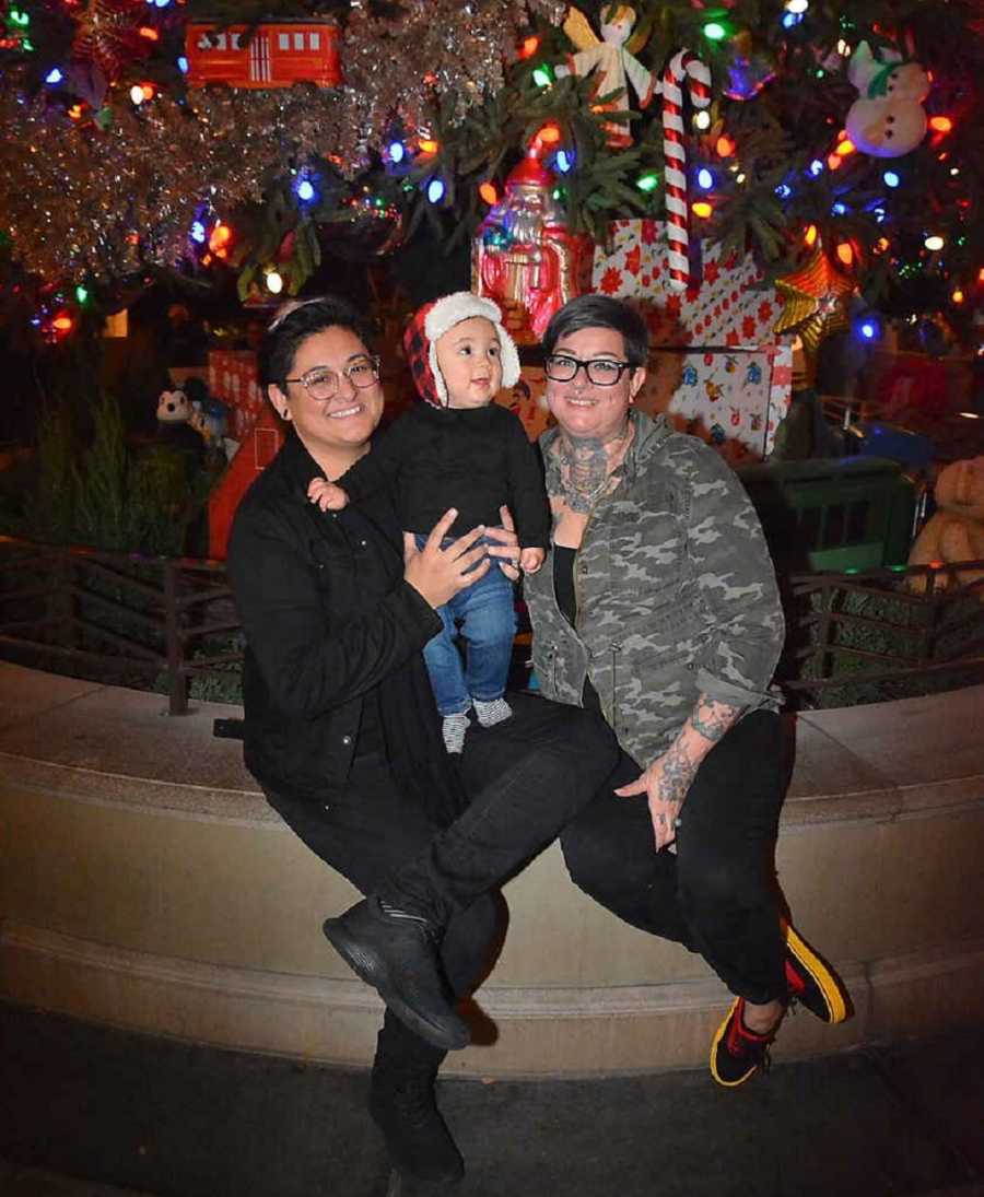 Mothers sit on ledge outside with their young son in front of Christmas tree