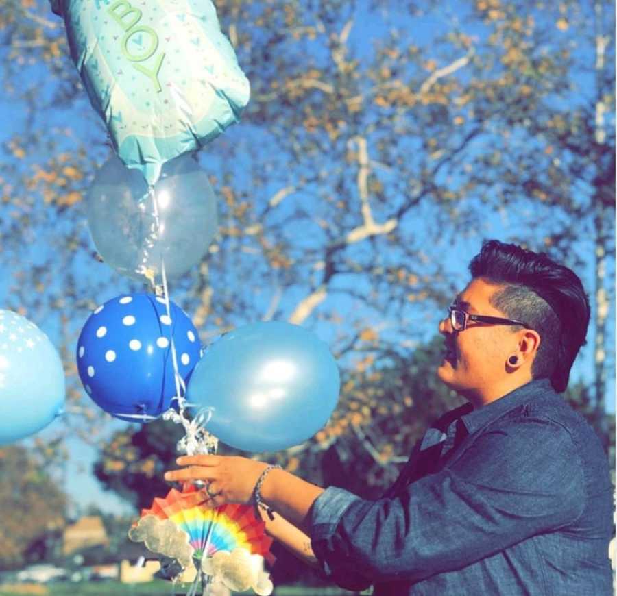 Woman stands outside holding strand of blue balloons with rainbow attached to end of it