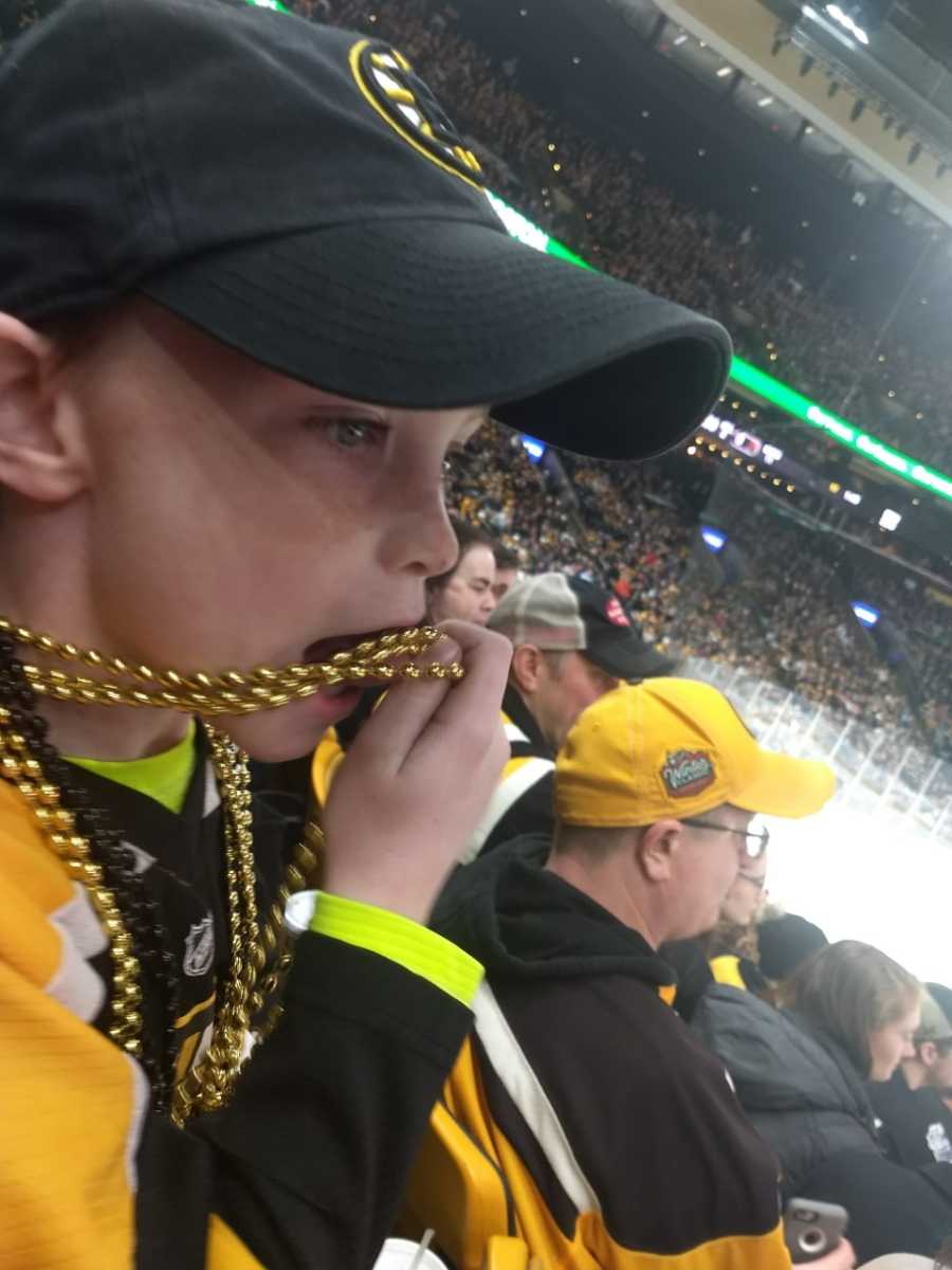 Little boy sits in Boston Bruins arena holding gold beads that are around his neck to his mouth