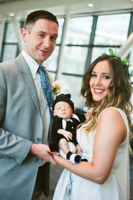 Bride and groom stand smiling as they hold newborn 