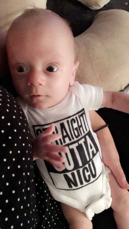 Newborn laying in woman's arms wearing onesie that says, "Straight outta NICU"