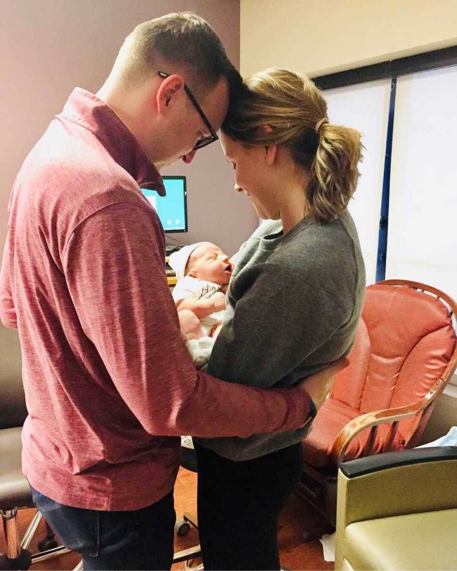 Husband stands holding wife's waist as she holds their adopted newborn baby in hospital room