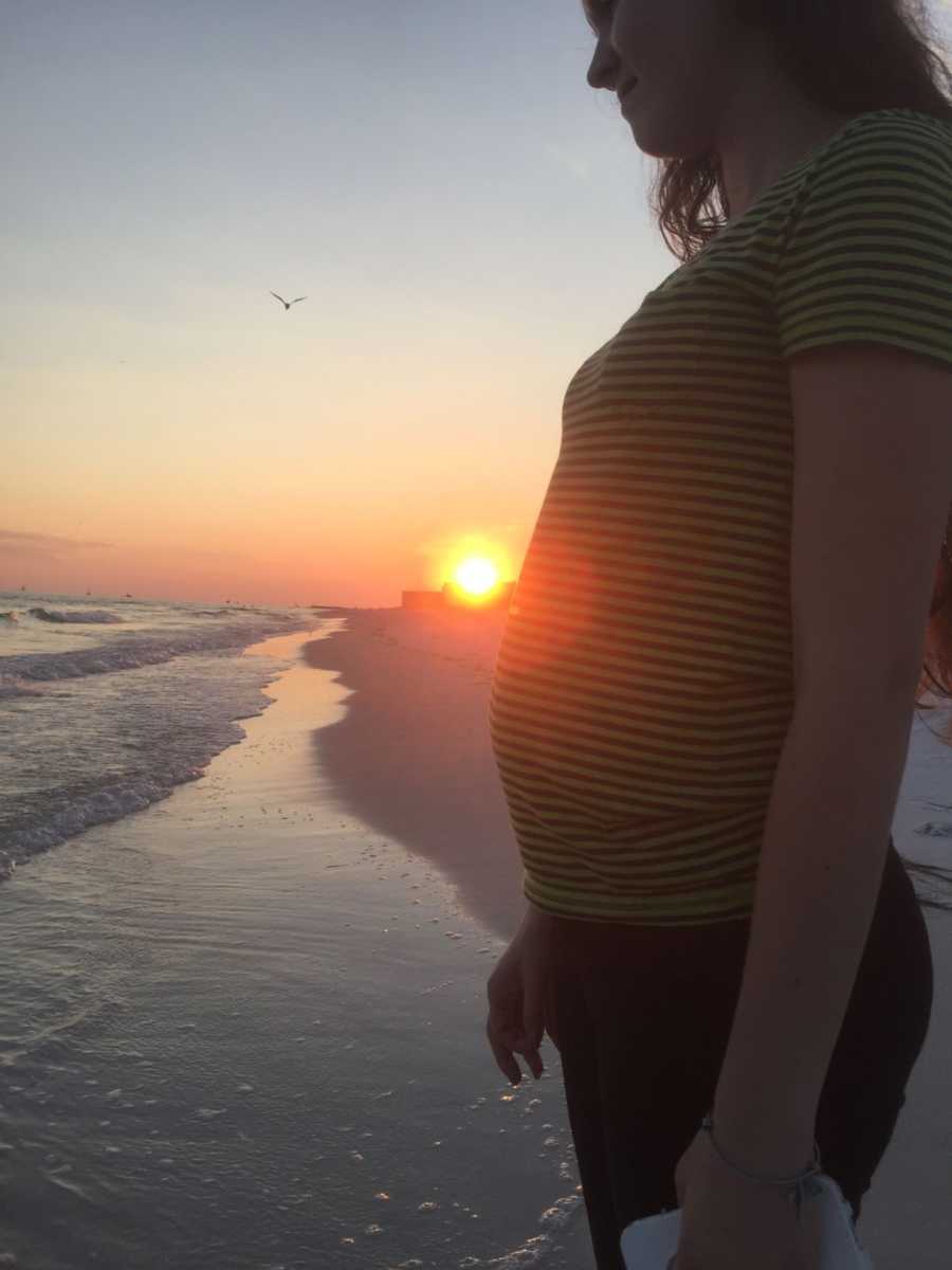 Pregnant woman stands on beach with sunset in background