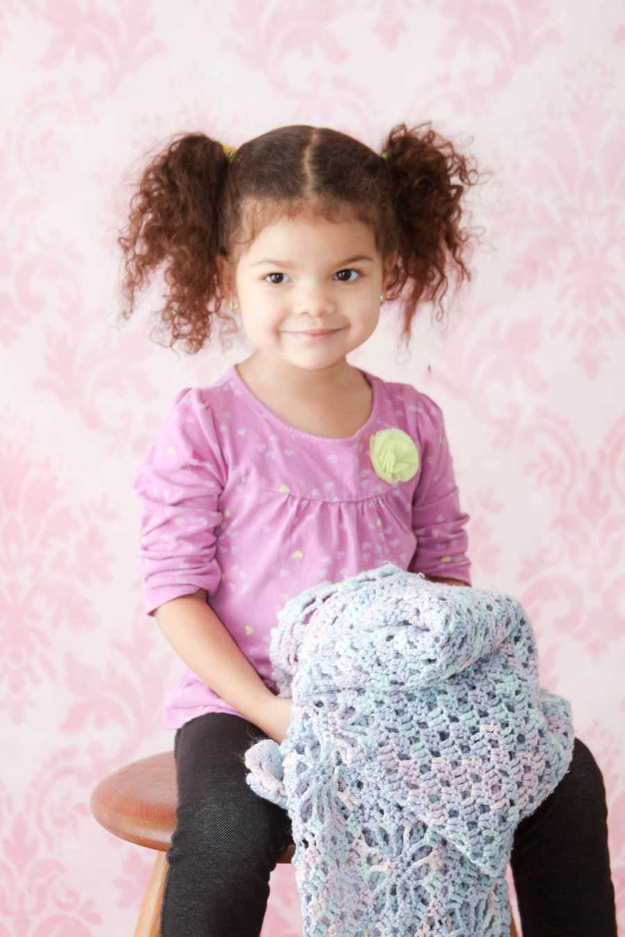 Little girl sits on wooden stool smiling as she holds knit blanket her great grandmother made her