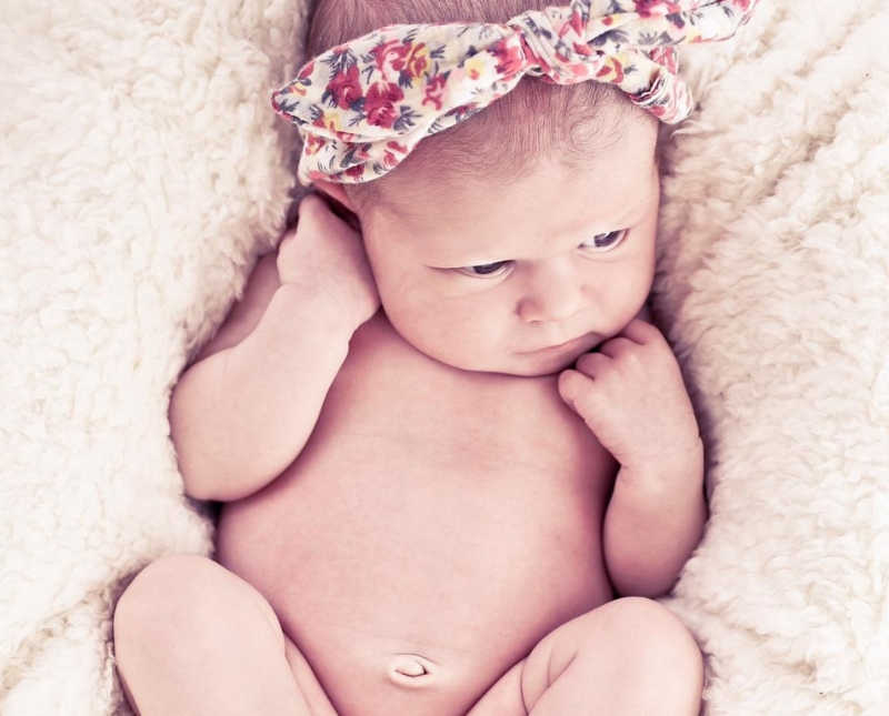 Baby with two mothers lay on back on white fuzzy blanket wearing floral headband