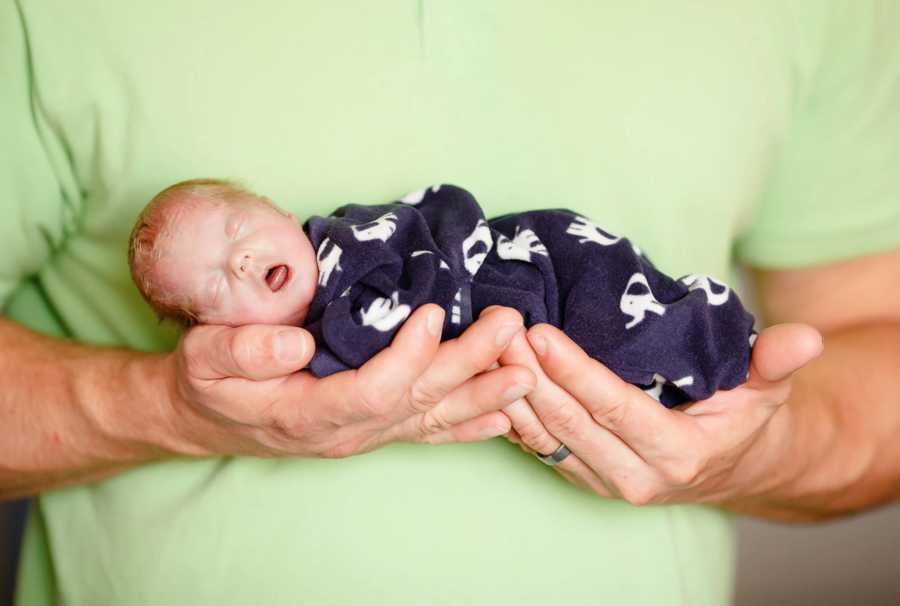 Preemie baby who didn't live long lays asleep in fathers hands in blue onesie with elephants 