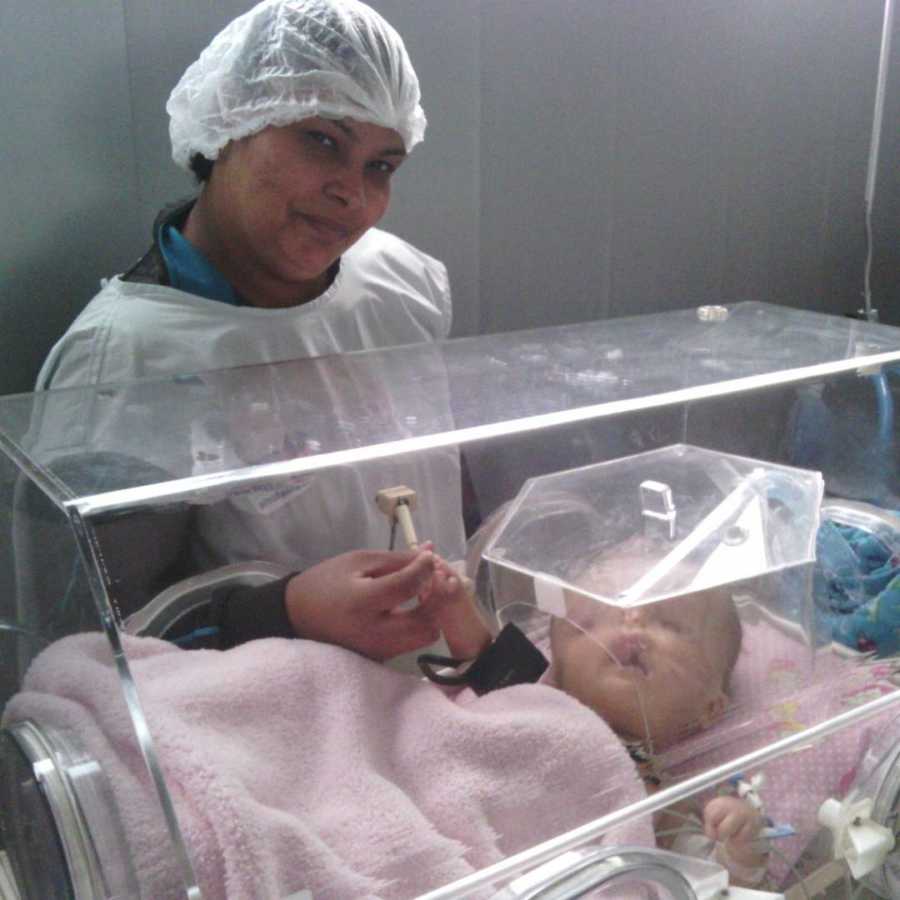Mother stands smiling beside newborn with birth defects in NICU holding baby's hand