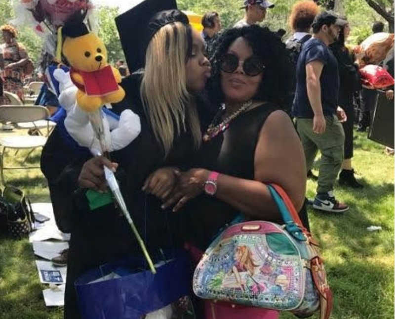 Young woman in cap and gown stands outside beside her mother as she leans in to kiss her cheek