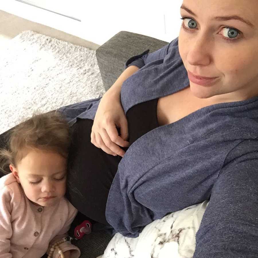 Pregnant woman smiles in selfie and she sits on couch and daughter lays asleep on her lap