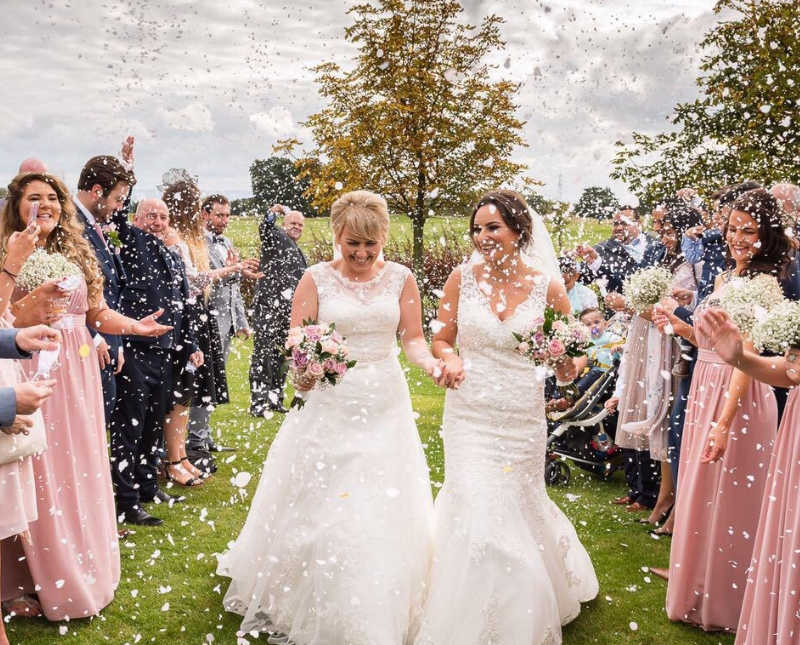 Brides walk hand in hand outside while guests of wedding shower them in white confetti