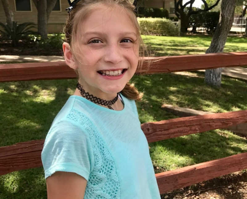 Young girl stands smiling outside beside wooden fence before leukemia diagnoses 