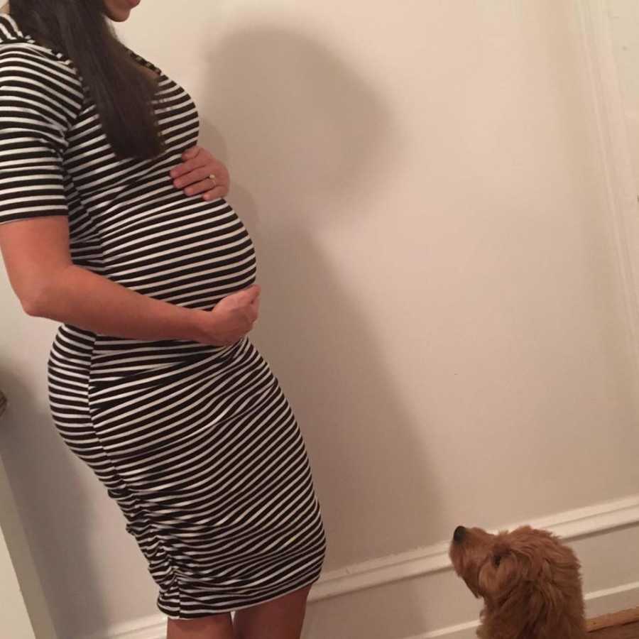 Pregnant woman stands in home holding her stomach as puppy looks up at her
