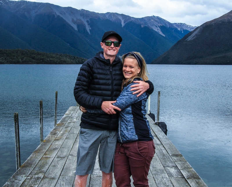 New Zealand track runner with Trigeminal neuralgia stands smiling on dock with girl friend with body of water and mountains in background