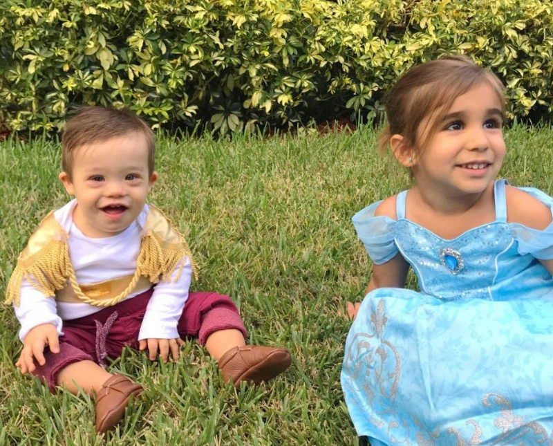 Little boy with down syndrome sits in grass dressed as prince charming besides sister who is dressed as Cinderella