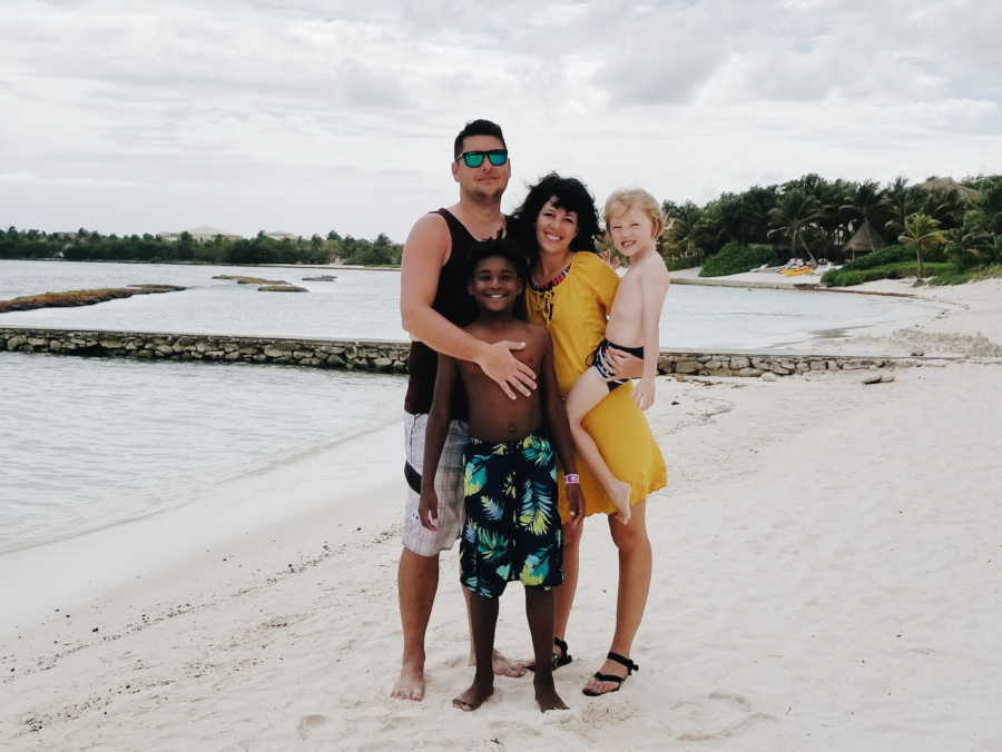 Husband and wife stand on beach while mother holds youngest son and older adopted son stands in front of them