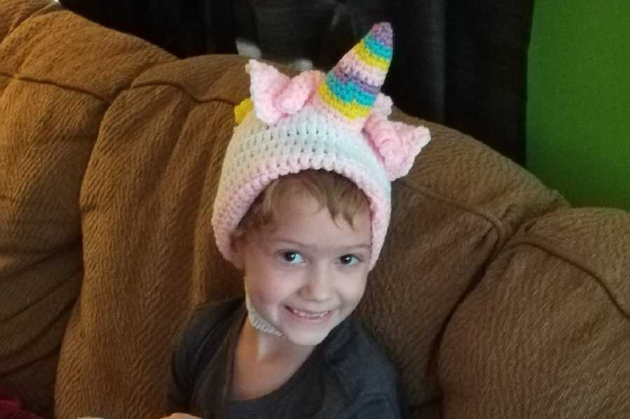 Little girl with brain cancer sits on couch smiling with knit unicorn hat on
