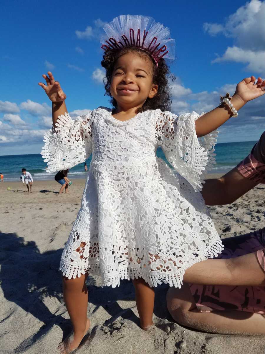 Little girl with prosthetic eyes stands on beach in white dress and birthday crown on