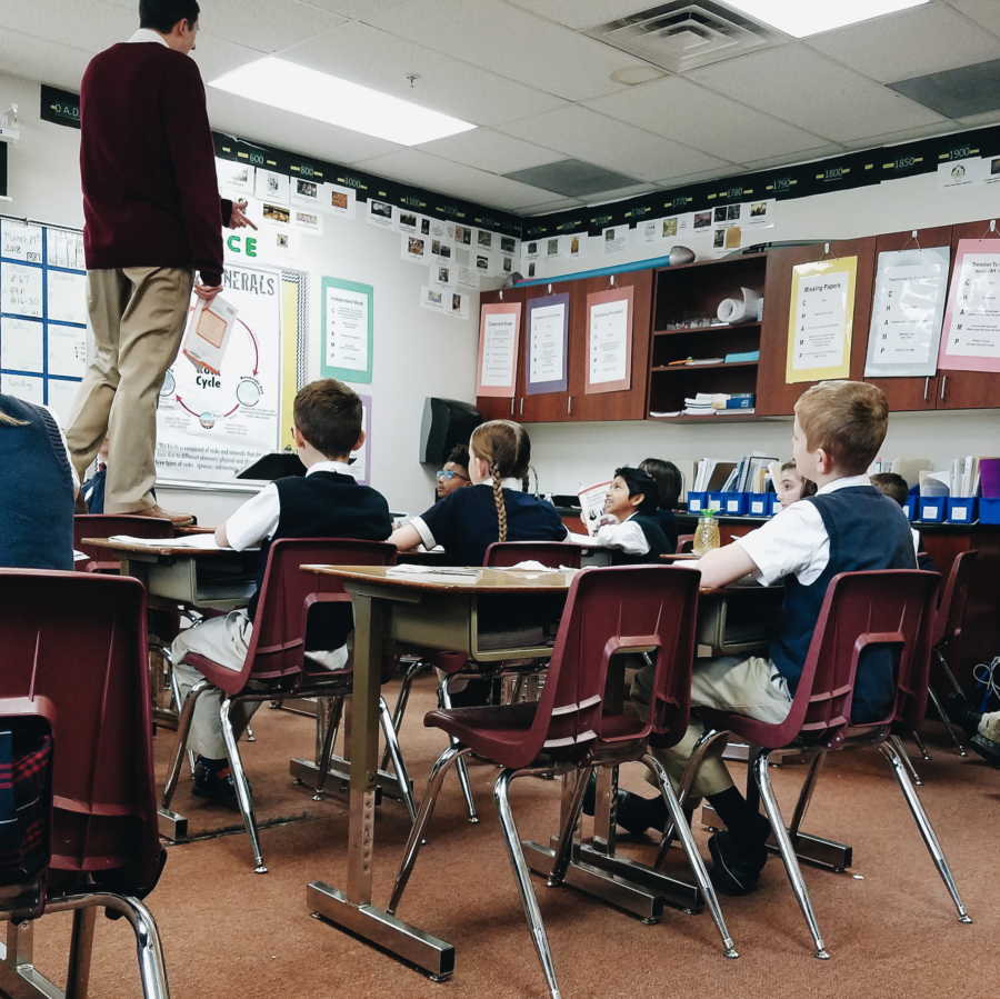 Teacher stands on desk in classroom while students look up at him