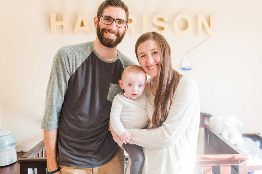 Husband and wife stand smiling with their baby in front of his crib