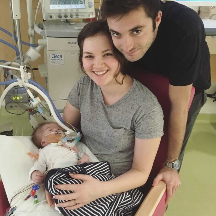 Mother sits smiling in chair holding baby with Hypoplastic Left Heart Syndrome while husband stands smiling behind her