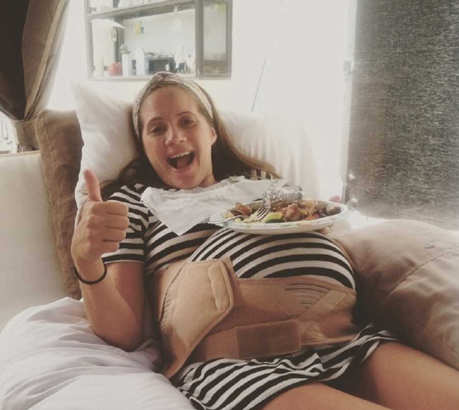 Woman pregnant with triplets lays on back with plate of food resting on her stomach as she holds a thumb up