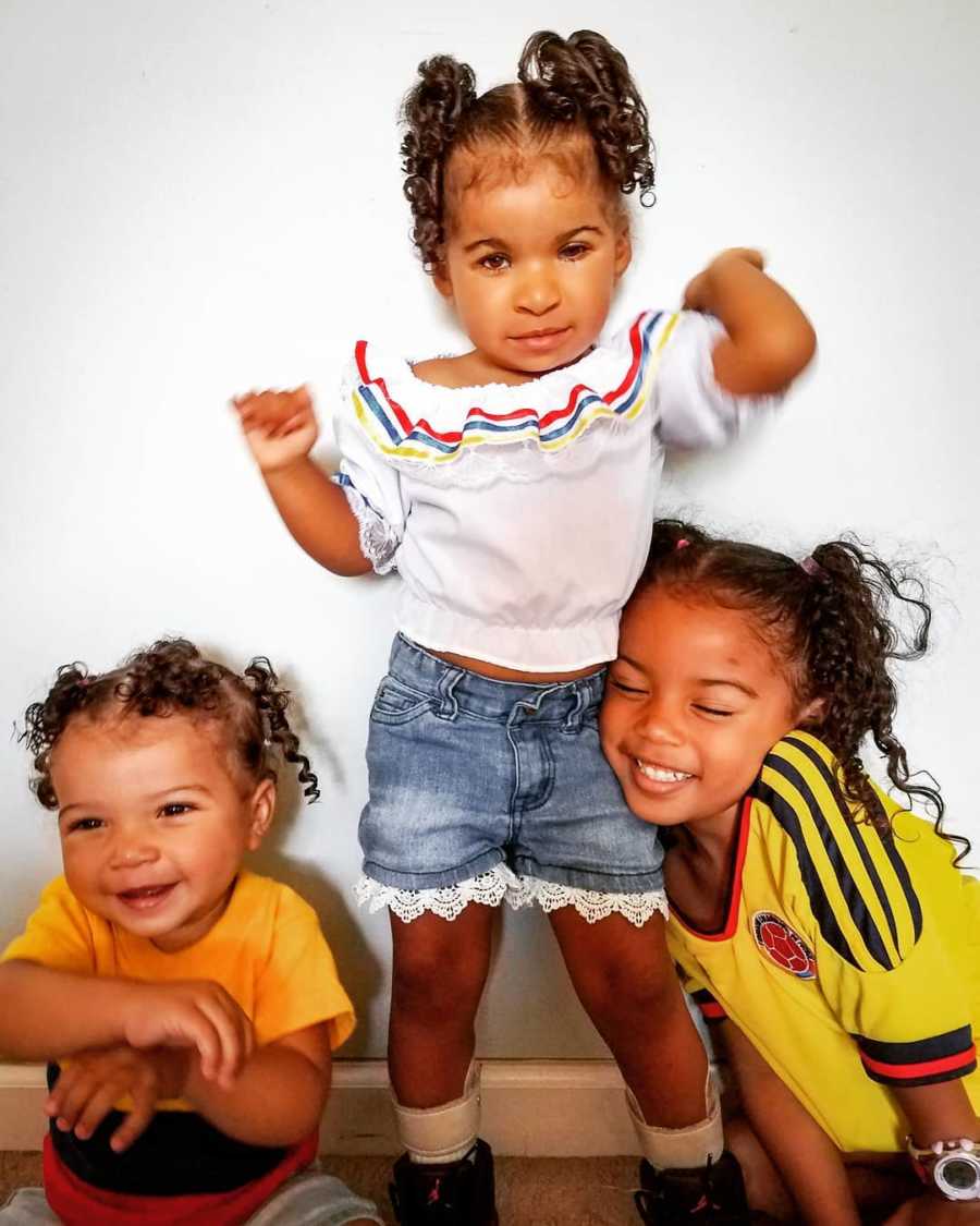 Little girl with prosthetic eyes stands in home while her older and younger sister sit on either side of her