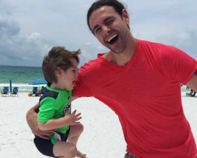 Man who has since passed stands on beach smiling as he holds son