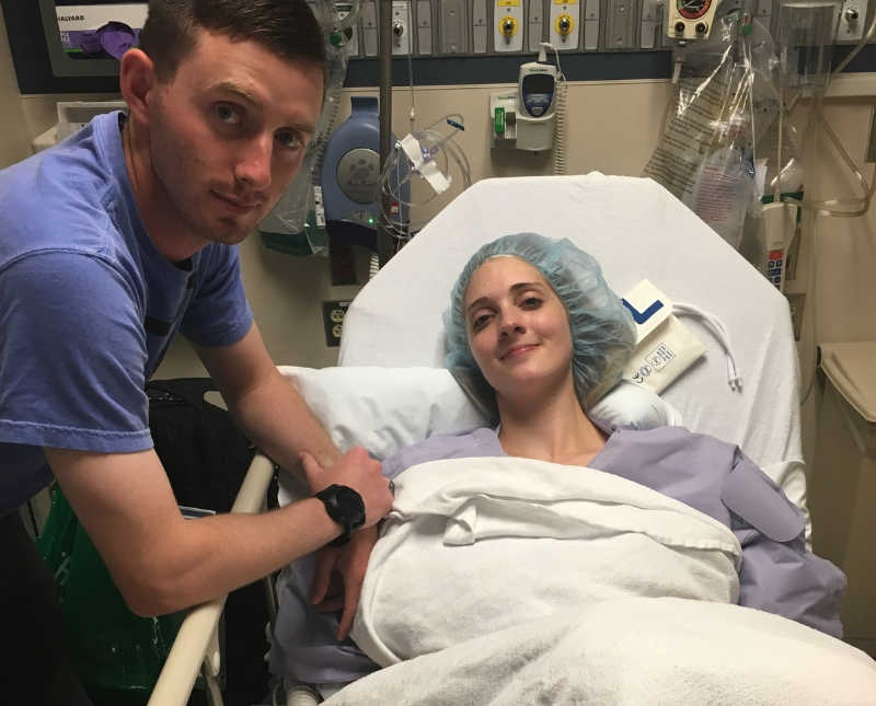 Woman lays in hospital bed after surgery that removed part of her colon with husband beside her