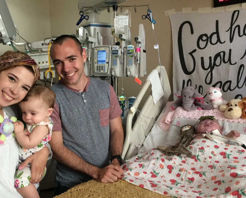 Husband and wife who holds one twin stand beside hospital bed where other twin with heart issues lies