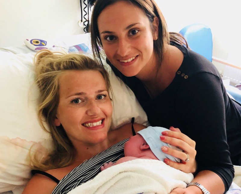 Woman smiles in hospital bed as she holds newborn while her wife leans over her