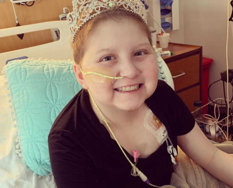 Intubated young girl sits in hospital bed smiling with crown on