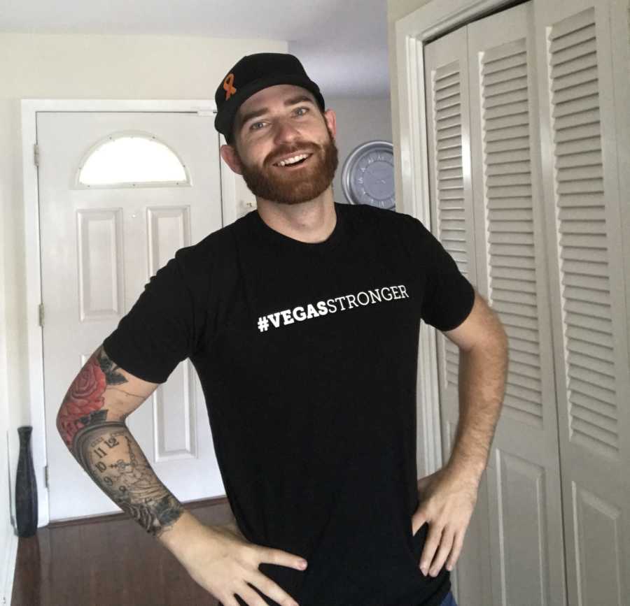 Man smiles in home with hands on his hips wearing shirt that says, #vegasstronger after surviving shooting