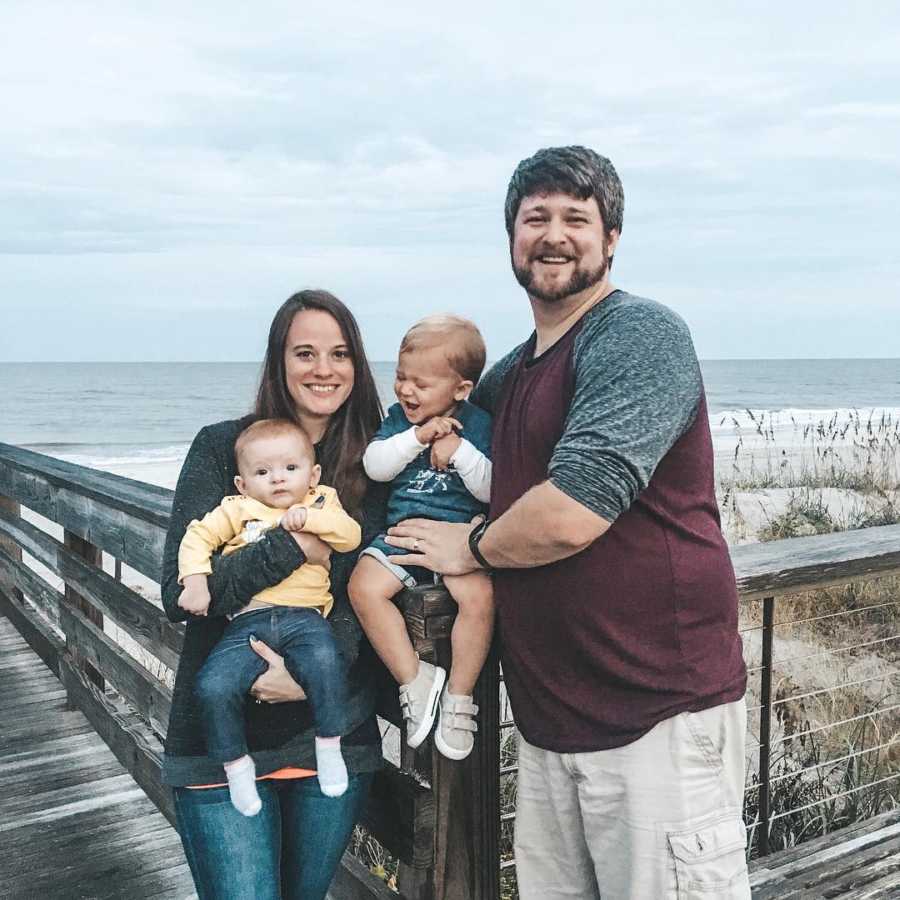 Husband and wife stand on boardwalk near beach smiling as they hold their two kids